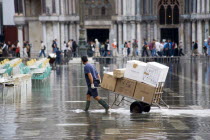 Aqua Alta High Water flooding in St Marks Square with a delivery man pulling a trolley of goods through the water with St Marks Basilica at the end of the flooded piazza