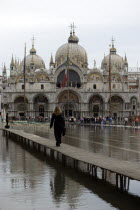 Aqua Alta High Water flooding in St Marks Square showing St Marks Basilica at the end of the flooded piazza and a resident walking to work on an elevated walkway