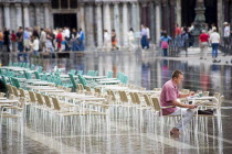 Aqua Alta High Water flooding in St Marks Square showing St Marks Basilica at the end of the flooded piazza with an artist painting a watercolour seated at a table in the water