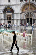 Aqua Alta High Water flooding in St Marks Square showing St Marks Basilica at the end of the flooded piazza with an artist painting a watercolour seated at a table in the water and local female reside...