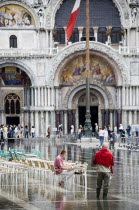 Aqua Alta High Water flooding in St Marks Square showing St Marks Basilica at the end of the flooded piazza with an artist painting a watercolour seated at a table in the water