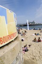 Tourists on the sand of the West Beach beside the pier. A sign reading Bournemouth is in the foreground