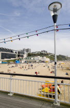 The East Beach from the pier with seafront restaurants and bars. Adults and children play in the sand on the beach and at the waters edge. Hotels and flats line the clifftop into the distance