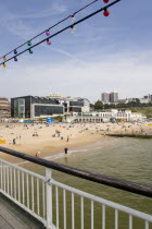 The East Beach from the pier with seafront restaurants and bars. Adults and children play in the sand on the beach and at the waters edge between the groynes. Hotels and flats line the clifftop into t...