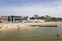 The East Beach with seafront restaurants and bars. Adults and children play in the sand on the beach and at the waters edge between the groynes. Hotels and flats line the clifftop into the distance