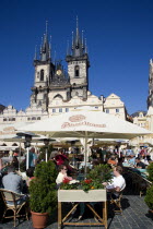 People sitting at restaurant tables under umbrellas in the Old Square in front of the Church of Our Lady before Tyn