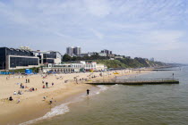 The East Beach showing the Imax Complex and seafront attractions with people on the beach and at the waters edge between the groynes. Clifftop hotels and flats in the distance