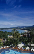 View over hotel swimming pool with tourists on sun loungers beside it towards curving sandy beach and distant tree covered hillside.