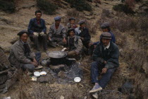 Group of labourers having lunch  sitting on ground around cooking pot supported by stones over fire.