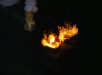 Gloved hand holding pole pouring molten gold into flaming mould