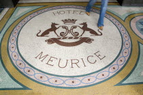 Woman walking across a mosaic with a crest and the words Hotel Meurice on the pavement outside the entrance door to the five star hotel in the Rue de RivoliFrench Western Europe European