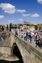 Tourists on the Charles Bridge across the Vltava River with Prague Castle and St Vitus Cathedral on the horizon and light clouds in the skyPraha Ceska Eastern Europe European Blue Religion Religious...