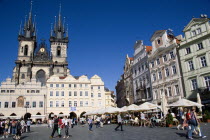 The Old Town Square with the Church of Our Lady before Tyn. Tourists sit at tables under umbrellas outside cafes and restaurants whilst others stroll in the square. Praha Ceska Eastern Europe Europea...