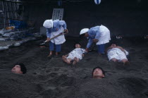 Sunabulo. Hot Sand Bath with people being covered in sand by two women leaving only their head showing above the surfaceAsia Asian Japanese Nihon Nippon