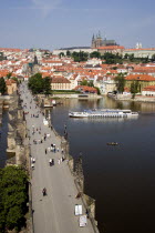 People walking on The Charles Bridge across the Vltava River leading to the Little Quarter. A river cruise boat moves up river to pass under the bridge with Prague Castle and St Vituss Cathedral on th...