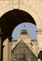 The French Tricolour flag flying from the centre of the Sully wing of the Musee du Louvre seen through the Arc de Triomph du Carrousel with the museum Pyramid entrance between them.Center European We...