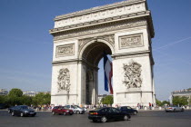 Tourists at the base of the Arc de Triomphe in Place Charles de Gaulle with traffic going around it. A large French Triclour flag hangs in the central arch of the ArcEuropean Western Europe