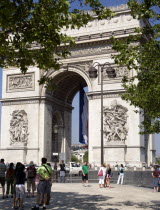 Tourists stand under trees looking at the Arc de Triomphe in Place Charles de Gaulle. A large French Triclour flag hangs in the central arch of the ArcEuropean Western Europe