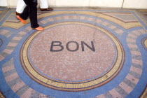 Pedestrians walk across a mosaic on the pavement with the word Bon at the centreCenter European French Western Europe