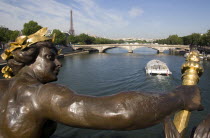 Bronze female figure holding a flame on the Pont Alexandre III bridge across the River Seine below with a passing Bateaux Mouches pleasure boat on the water and the Eiffel Tower beyondEuropean French...