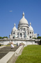 Montmartre The Facade of the church of Sacre Couer with tourists by the ballistrade at the front looking over Paris and others walking up the steps leading to the church. A plane flies in the sky abov...