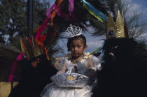 Little girl dressed in white flanked by two masked attendants in costume on float during carnival.