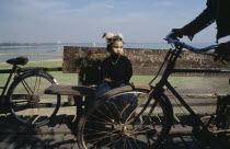 Young girl with face decorated with leaf patterns sitting on wooden bench on U-Bein bridge with wheels of passing cyclist in the foreground.Burma Myanmar Asian Burmese Kids Southeast Asia  Burma Mya...