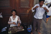 Women cooking meat skewers on barbecue with man eating nearby