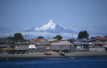 Coastal village buildings near Chiloe from sea with snow capped volcanic peak wreathed in cloud behind.American Chilean Hispanic Latin America Latino Scenic South America