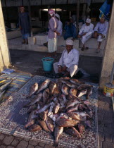 Mutrah fish market.  Vendor sitting behind display of whole fish scattered with ice laid out on matting on ground in front of him.  Group of men sitting behind. Male Man Guy Middle East Omani United...