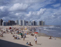 Busy  sandy beach with people sunbathing  playing ball games and in surf.  Overlooked by line of high rise buildings.  American Beaches Hispanic Latin America Latino Resort Seaside Shore South Americ...