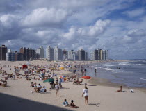 Busy  sandy beach with people sunbathing  playing ball games and in surf.  Overlooked by line of high rise buildings.  American Beaches Hispanic Latin America Latino Resort Seaside Shore South Americ...
