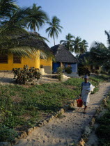 The Tides Hotel.  Brightly painted thatched huts amongst palm trees with maid carrying cleaning equipment and fresh towels on path in foreground.chambermaid tourism African Eastern Africa Tanzanian C...