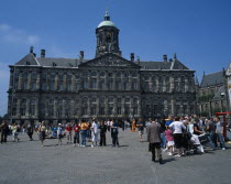 Damm Square.  Royal Palace exterior facade with domed clock tower and weather vane  pediment with relief carving and multiple windows.  Crowds in foreground.North Seventeenth Century Dutch classicism...