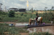 Women washing clothes in foreground with oil refinery behind.African Ecology Entorno Environmental Environnement Green Issues Nigerian Western Africa Female Woman Girl Lady Female Women Girl Lady Cle...