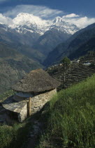 Sanctuary Trek. Circular stone building with thatched roof overlooking sloping agricultural terracing. Snow capped mountains Annapurna South and Hiunchuli in the backgroundAsia Asian Nepalese Scenic...