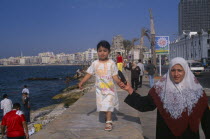 Corniche Waterfront. A mother wearing an Islamic headscarf holding her daughters hand while she walks along wall above her.African Middle East North Africa Kids Mum Religious Religion