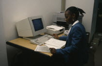 Smartly dressed woman working on computer at office desk with printer  telephone and photo copier.African Botswanan Female Women Girl Lady Southern Africa Female Woman Girl Lady