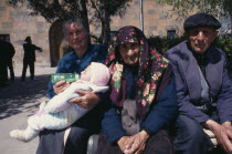Woman bottle feeding baby beside elderly couple sitting in sunshine.Etchmiadzin Armenian Asia Asian European Kids Middle East Babies Female Women Girl Lady Old Senior Aged Female Woman Girl Lady