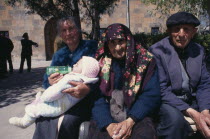 Woman bottle feeding baby beside elderly couple sitting in sunshine.Etchmiadzin Armenian Asia Asian European Kids Middle East Babies Female Women Girl Lady Old Senior Aged Female Woman Girl Lady