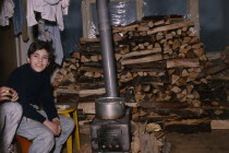 Boy sitting beside wood burning stove in house with large stack of wood behind during winter time.cold Armenian Asia Asian European Kids Middle East