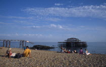 Groups of people sitting on the beach by the ruined West Pier in late afternoon sunshine.European Great Britain Northern Europe UK United Kingdom British Isles Beaches Kids Resort Sand Sandy Seaside...