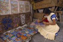 Young woman painting design on piece of bark with masks and other paintings displayed beside her.