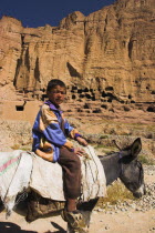 Boy on donkey infront of caves in cliffs near empty niche where the famous carved small Budda once stood 180 foot high before being destroyed by the Taliban in 2001
