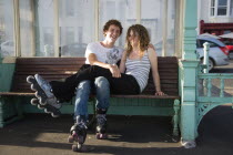 Young couple sat on bench on the seafront promenade.One wearing Rollerblades  one wearing RollerbootsRollerblading  Skates Skating European Great Britain Northern Europe UK United Kingdom