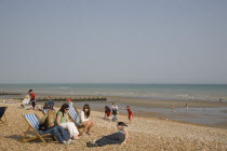 A group of girls sitting on deck chairs on shingle beach with children playing behind
