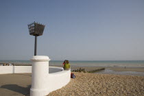 Two young women sitting on a white wall facing each other talking near a torch beacon. Sand and shingle beach with families seen at the waters edge behind