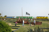 Families enjoying pitch and putt mini golf games in Norfolk Gardens next to seafront. A skull and cross bones flag flying. Colourful beach huts behind.
