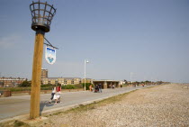 People walking along the promenade next to shingle beach with a torch beacon in the foreground