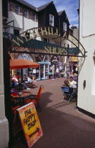 Westhill Arcade. Gate leading to shops with people sitting at cafe tables. A sign advertising The Happy Shop in the foreground.